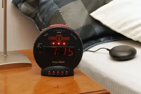 A Sonic Bomb alarm clock on a nightstand showing 7:15, with its bed shaker on the mattress beside it