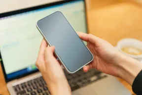 Two hands holding a phone with a dark screen in front of an open laptop