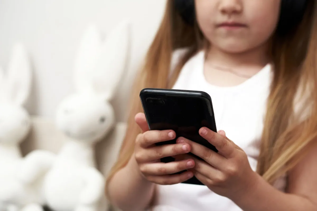 A young kid holding a black phone in both hands, with blurred plush rabbits behind