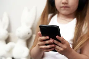 A young kid holding a black phone in both hands, with blurred plush rabbits behind
