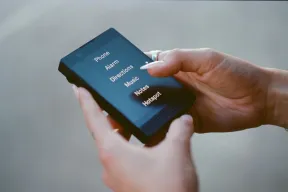 A hand tapping a minimalist dumbphone whose text-only screen lists tools like Alarm, Directions, and Hotspot