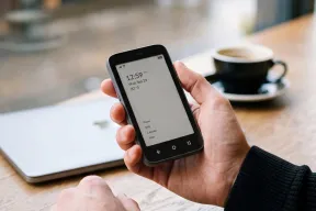 A hand holding a small E Ink phone with a plain home screen, next to a coffee cup and a laptop