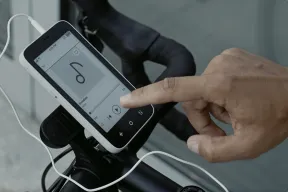 A hand tapping play on an E Ink phone showing a music player, mounted on a bike handlebar with wired earbuds