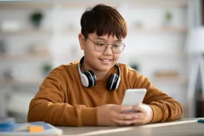 A smiling kid in glasses, with headphones around the neck, looks at a phone held at a desk