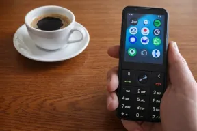 A hand holding the Dumber Mini keypad phone, its small screen showing app icons, beside a coffee cup