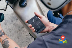 A motorcycle rider holds a dumbphone showing the next turn on a simple map, beside a map pin icon