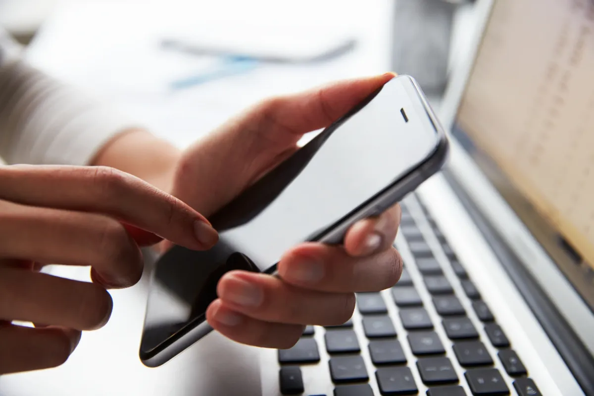 Two hands holding a phone with a blank screen above a laptop keyboard