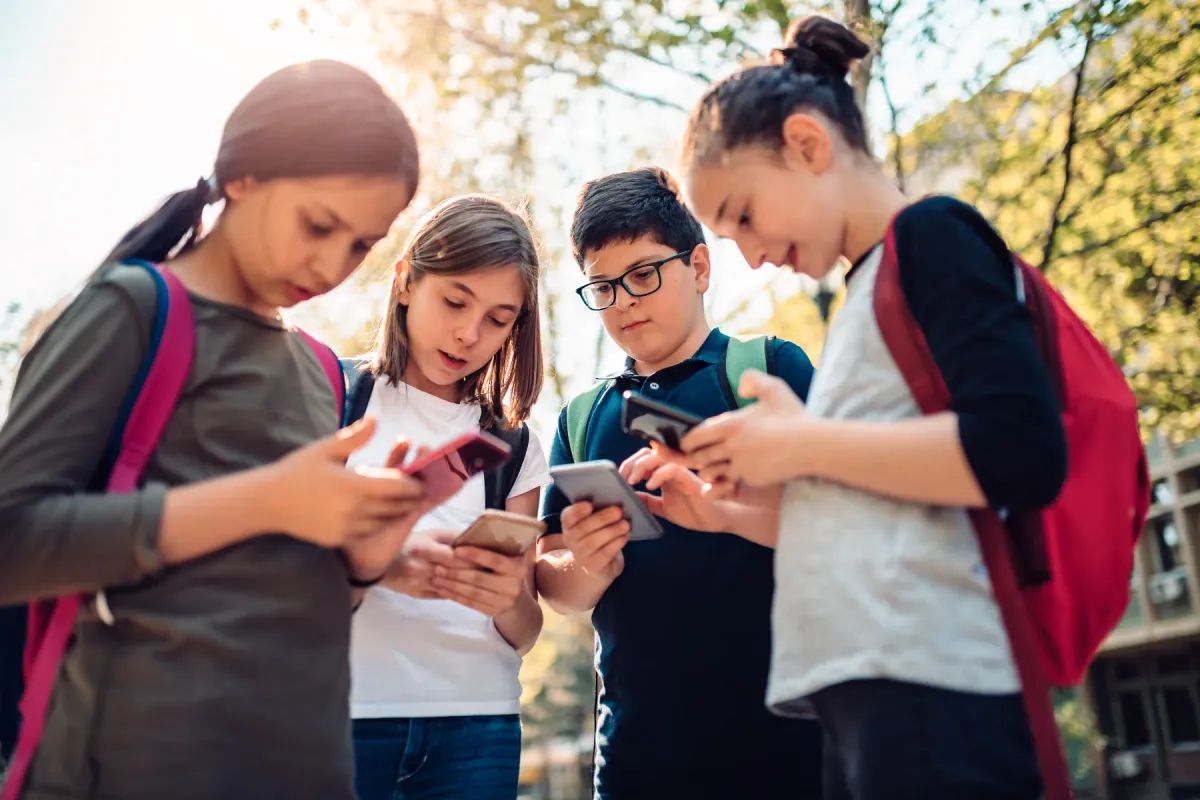Four kids with school backpacks outdoors, each child looking at a phone