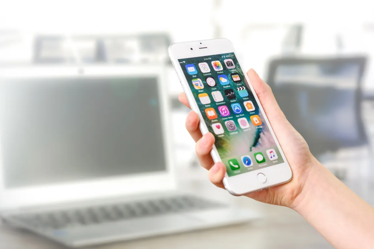 A hand holding an iPhone with a full Home Screen of apps, in front of a laptop on a desk